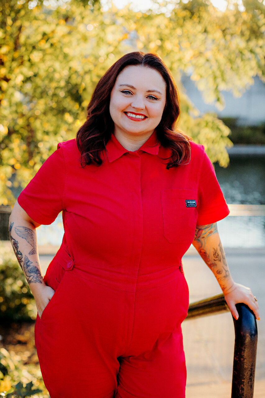 white woman standing in front of a tree. She is wearing a red jumper, bright read lipstick and had aubrun shoulder length hair parted down the middle.